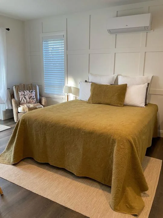 Second Cozy bedroom featuring a mustard-colored bedspread, white pillows, paneled accent walls, a wicker chair with floral cushions, and natural lighting through a window with blinds Cozy bedroom featuring a mustard-colored bedspread, white pillows, paneled accent walls, a wicker chair with floral cushions, and natural lighting through a window with blinds